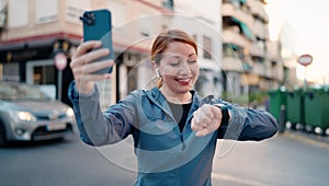 Young redhead woman wearing sportswear make selfie by smartphone looking stopwatch at street