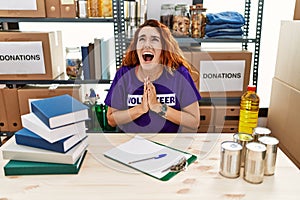 Young redhead woman wearing volunteer t shirt at donations stand begging and praying with hands together with hope expression on