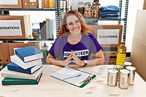 Young redhead woman wearing volunteer t shirt at donations stand hands together and fingers crossed smiling relaxed and cheerful