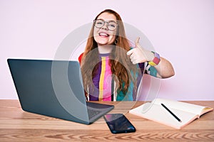 Young redhead woman working at the office with laptop smiling happy and positive, thumb up doing excellent and approval sign