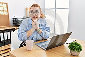 Young redhead woman working at the office using computer laptop laughing nervous and excited with hands on chin looking to the