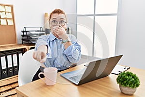 Young redhead woman working at the office using computer laptop laughing at you, pointing finger to the camera with hand over