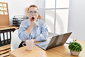 Young redhead woman working at the office using computer laptop looking fascinated with disbelief, surprise and amazed expression