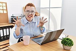 Young redhead woman working at the office using computer laptop smiling funny doing claw gesture as cat, aggressive and sexy