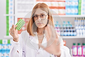 Young redhead woman working at pharmacy drugstore holding birth control pills with open hand doing stop sign with serious and