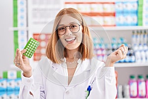 Young redhead woman working at pharmacy drugstore holding birth control pills screaming proud, celebrating victory and success