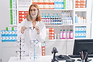 Young redhead woman working at pharmacy drugstore holding glasses smiling and laughing hard out loud because funny crazy joke