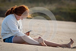 Young redheaded girl using a phone beside a river at the beach