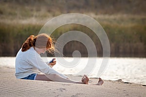 Young redheaded girl using a phone beside a river at the beach