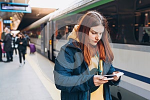 Young redhhead woman with backpack checking her ticket and using smartphone on background train. Railroad transport