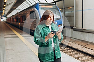 Young redhhead woman with passport waiting on station platform and using smart phone. Railroad transport concept. Travel