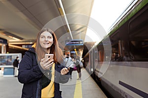 Young redhhead woman waiting on station platform with backpack on background electric train using smart phone. Railroad