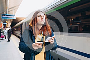Young redhhead woman waiting on station platform with backpack on background electric train using smart phone. Railroad