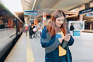 Young redhhead woman waiting on station platform with backpack on background electric train using smart phone. Railroad