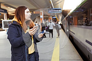 Young redhhead woman waiting on station platform with backpack on background electric train using smart phone. Railroad