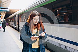 Young redhhead woman waiting on station platform with backpack on background electric train using smart phone. Railroad