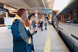 Young redhhead woman waiting on station platform with backpack on background electric train using smart phone. Railroad