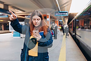 Young redhhead woman waiting on station platform with backpack and pointing finger on background train using smart phone