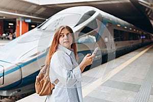 Young redhhead woman waiting train on station platform with backpack and using smart phone. Railroad transport concept