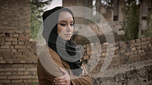 Young refugee muslim woman in hijab standing near ruined building and looking at camera with frightened and scared