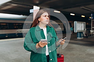 Young refugee redhhead woman crying and waiting train on station platform, she lost and using smart phone. Railroad