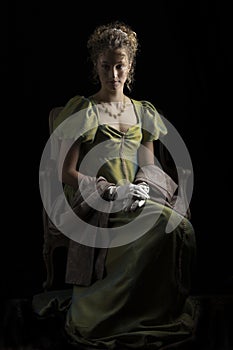 A young Regency woman wearing a green shot silk dress and sitting in front of a dark backdrop