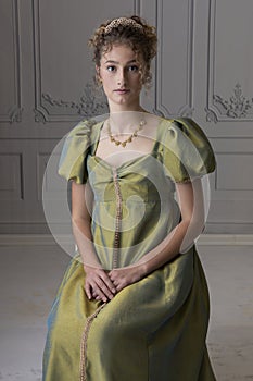 A young Regency woman wearing a green shot silk dress and sitting in front of a white paneled wall