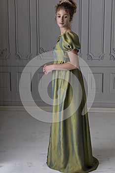 A young Regency woman wearing a green shot silk dress and standing in front of a white paneled wall