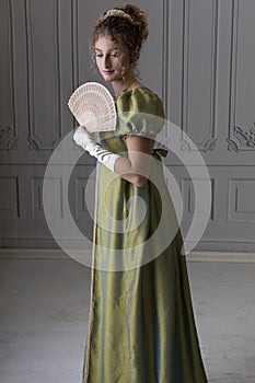 A young Regency woman wearing a green shot silk dress and standing in front of a white paneled wall and holding a fan