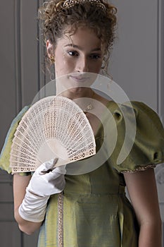 A young Regency woman wearing a green shot silk dress and standing in front of a white paneled wall and holding a fan