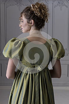 A young Regency woman wearing a green shot silk dress and standing in front of a white paneled wall shown in back view