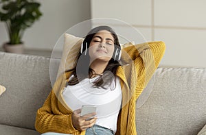 Young relaxed arab woman in wireless headphones relaxing on couch, listening to music at home, free space