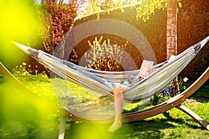 Young relaxed girl reading book in hammock in garden at home at bright sunset. Slow living,