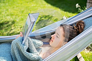 Young relaxed girl reading travel book in hammock in garden at home at bright sunny day. Slow living,
