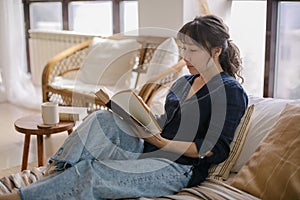 Young relaxed asian woman in casual outfit reading book and drinking tea while relaxing on sofa at home