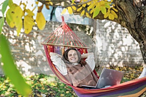 Young relaxed happy woman freelancer with laptop lying in hammock at backyard of country house on autumn day