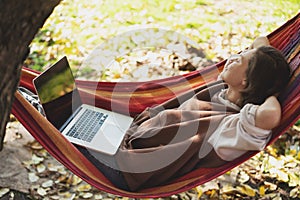 Young relaxed happy woman freelancer with laptop lying in hammock at backyard of country house on autumn day