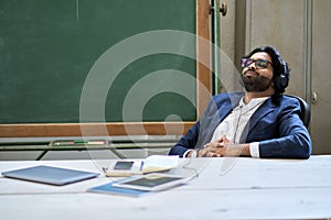 Young relaxed indian business man wearing headphones sitting listening to music.