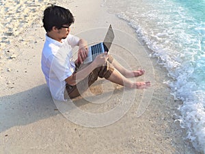 Young relaxed man with laptop sitting on the sandy beach with soft waves. Internet of things concept.