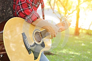 Young relaxed man in red shirt holding an acoustic guitar and playing music at the park outdoors with sunshine filters background.