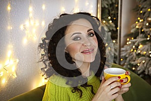 Young relaxed smiling woman sitting with coffee cup on sofa in morning after New Year party, happy female resting with mug of hot