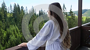 Young relaxed woman in bathrobe standing on hotel room balcony and looking on mountains and forest. Concept of tourism