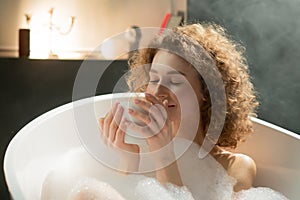 Young relaxed woman lying in the bath enjoys the smell of fragrant tea. Attractive woman lies in a bath with foam and