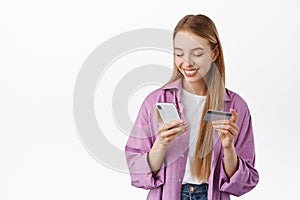 Young relaxed woman shopping online, paying with credit card and mobile phone. Girl looks at smartphone screen while