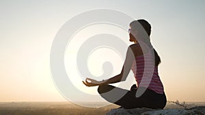 Young relaxed woman sitting outdoors on a big stone enjoying warm summer day. Girl meditating and relaxing on nature at sunset.