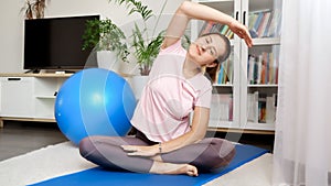 Young relaxed woman sitting on yoga mat at living room and stretching hand and back muscles