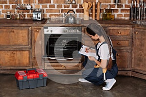 young repairwoman examining kitchen oven and writing