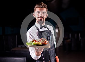 Young restaurant employee serves salad on a black background.