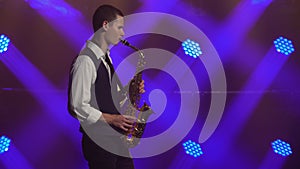 A young retro stylish guy plays on the golden shiny saxophone in the blue spotlights on stage. Dark studio with smoke