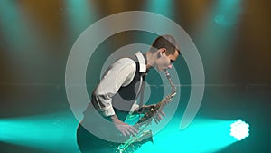 A young retro stylish guy plays on the golden shiny saxophone in the turquoise spotlights on stage. Dark studio with